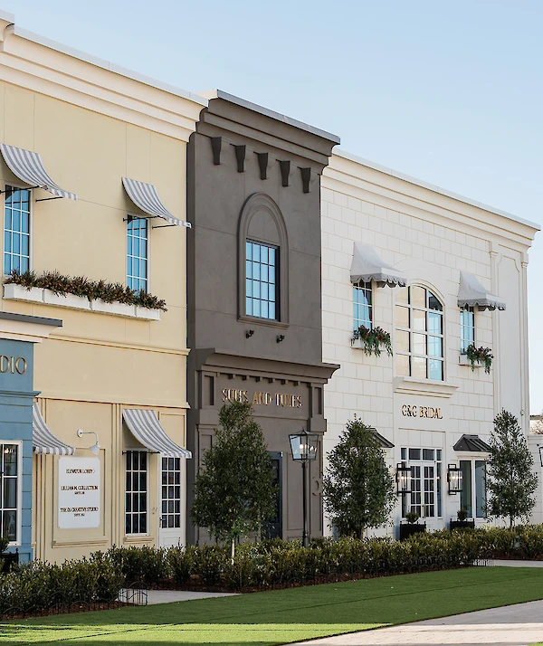 Row of stylish boutique storefronts with decorative awnings, tall windows, flower boxes, and manicured greenery, including shops labeled ‘Suits and Tuxes’ and ‘C&C Bridal’ under a clear blue sky.