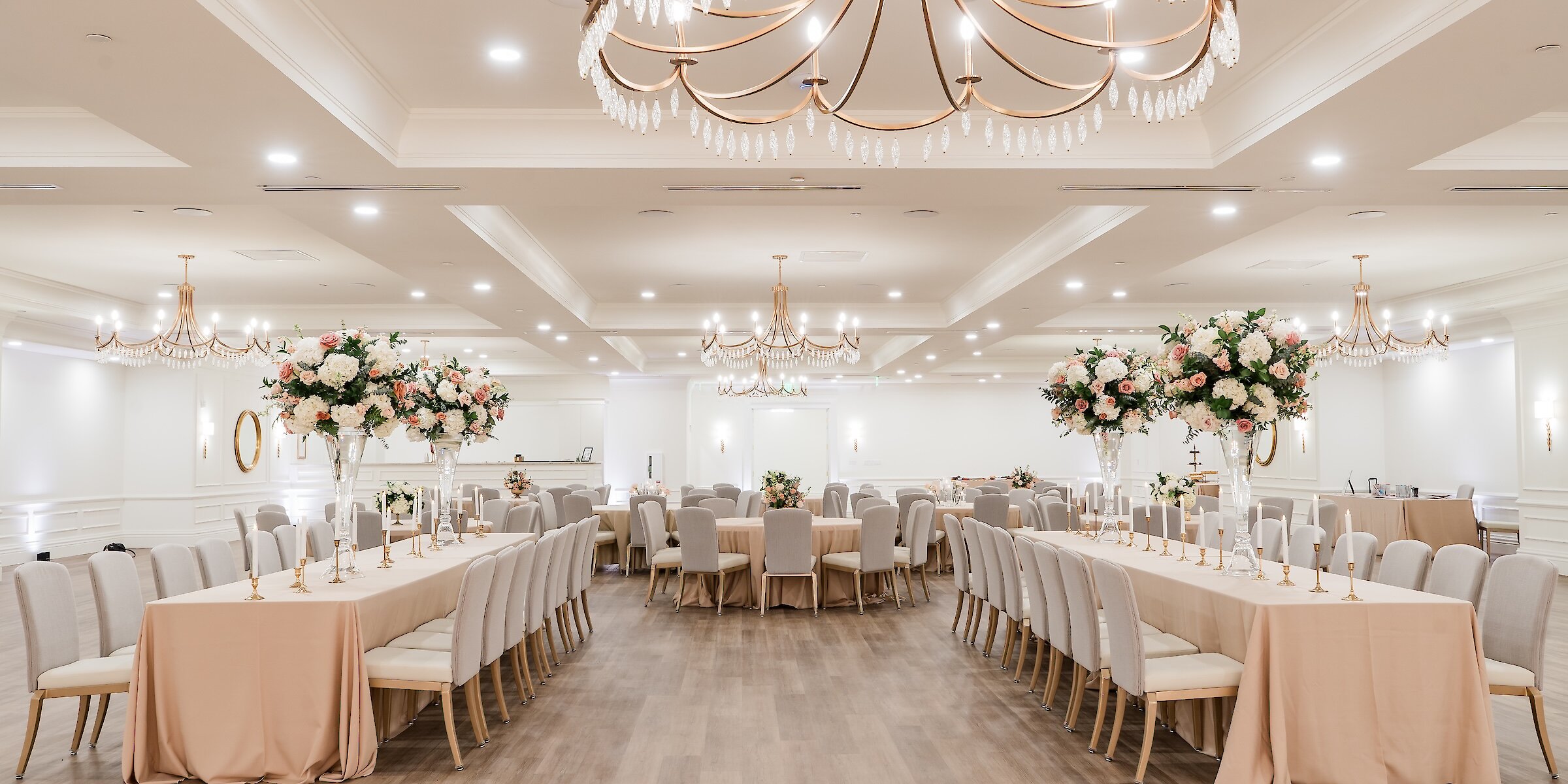 Elegant wedding reception hall with long tables, tall floral centerpieces, gold chandeliers, and neutral-toned décor arranged for a formal celebration