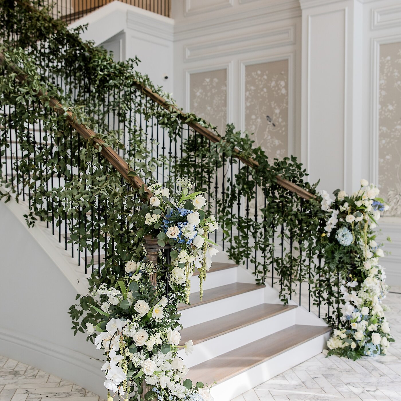Floral-decorated grand staircase adorned with lush greenery and white and blue blooms inside an elegant venue.