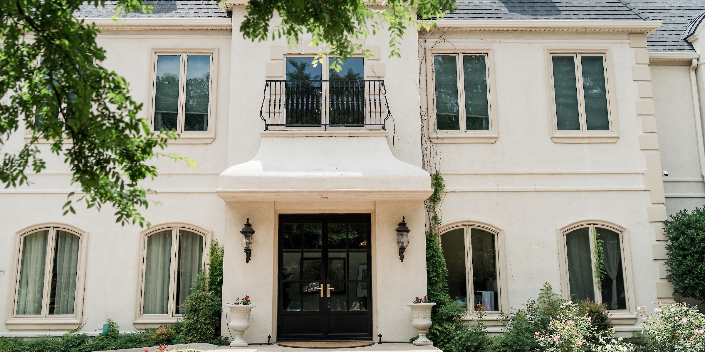 The symmetrical front exterior of a luxurious French-style mansion wedding venue featuring cream stucco, black doors, and a small balcony.
