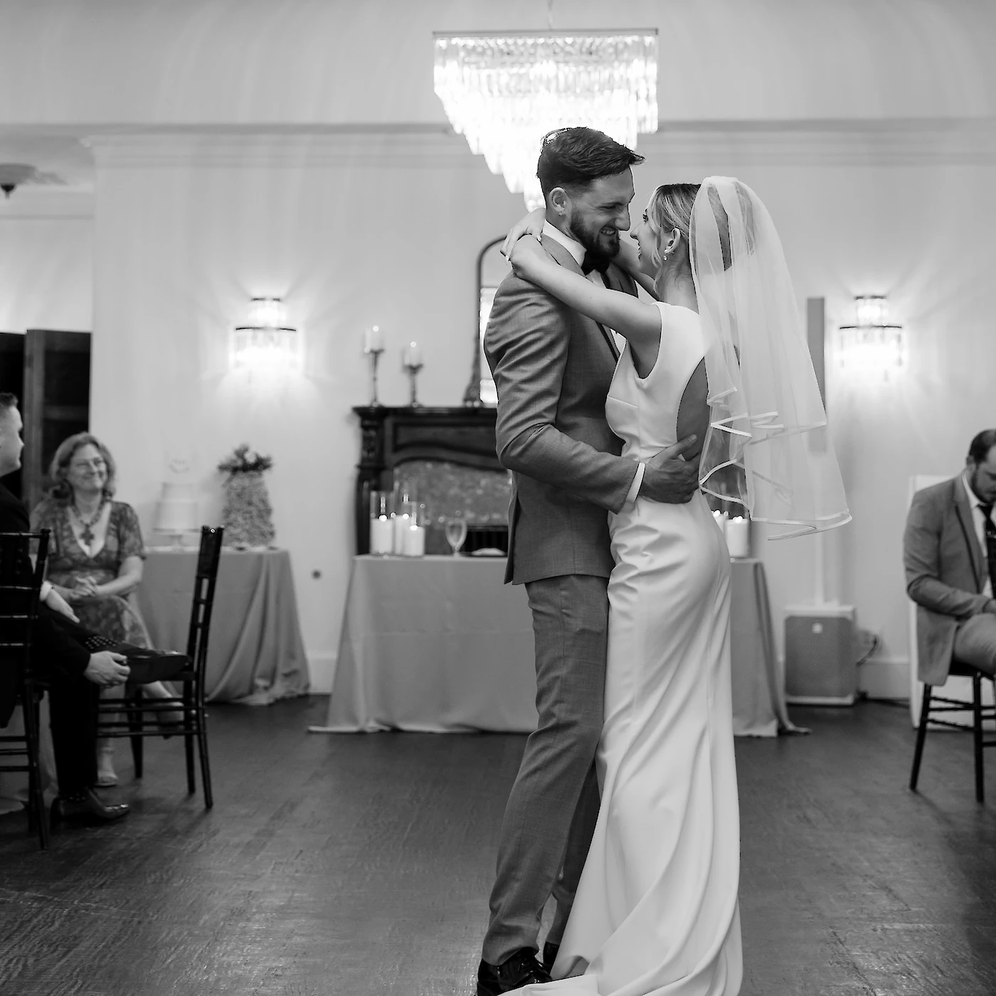 A black and white photo of the bride and groom sharing an intimate embrace during their first dance in a ballroom with a crystal chandelier.