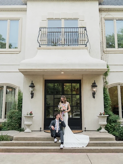 The bride and groom pose on the front steps of a white French mansion, with the groom sitting and kissing the bride's hand, holding a white and pink bouquet.