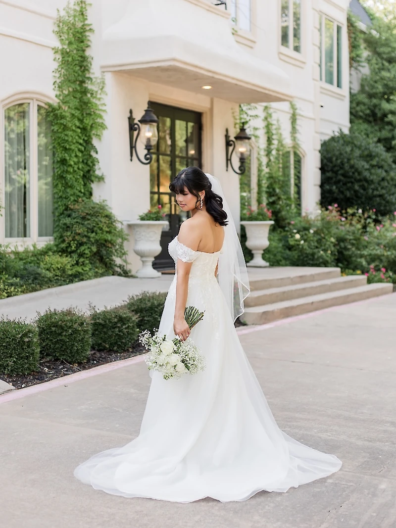 The bride in an off-the-shoulder gown holds a white baby's breath bouquet and looks over her shoulder, posing outside a grand white mansion.