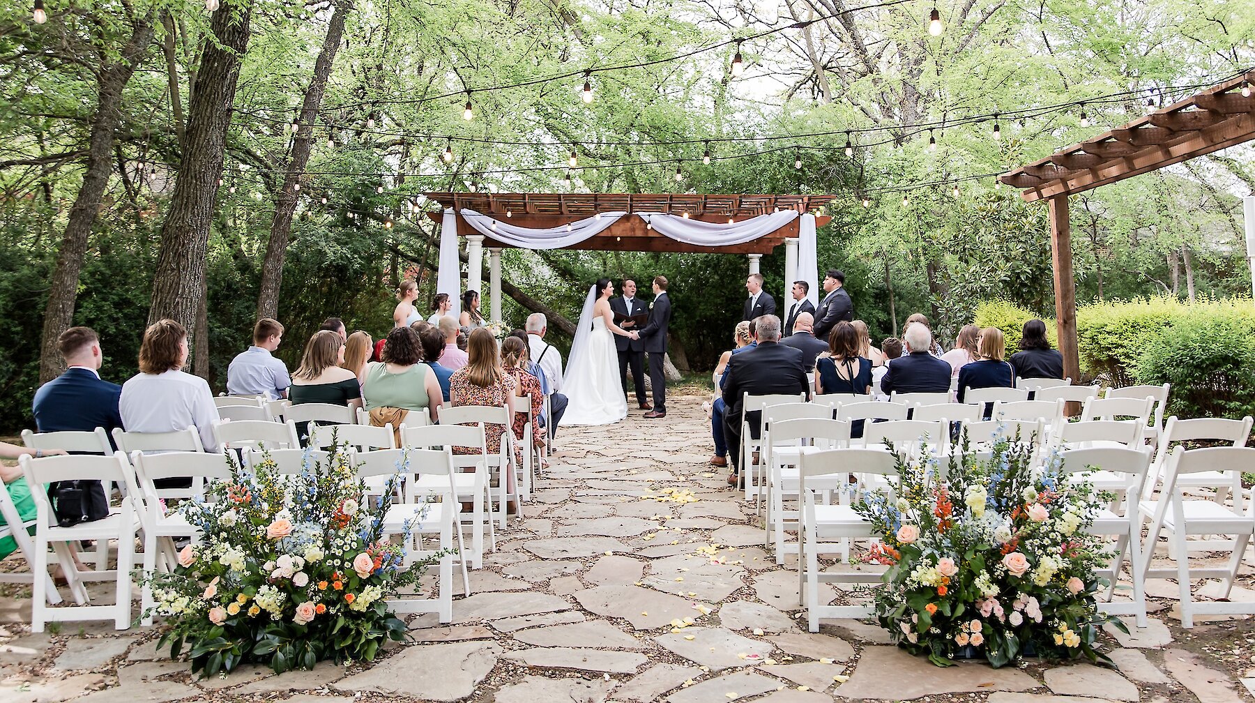 An outdoor wedding ceremony taking place under a wooden pergola with white drapery and string lights, with guests seated on white chairs on a flagstone path.