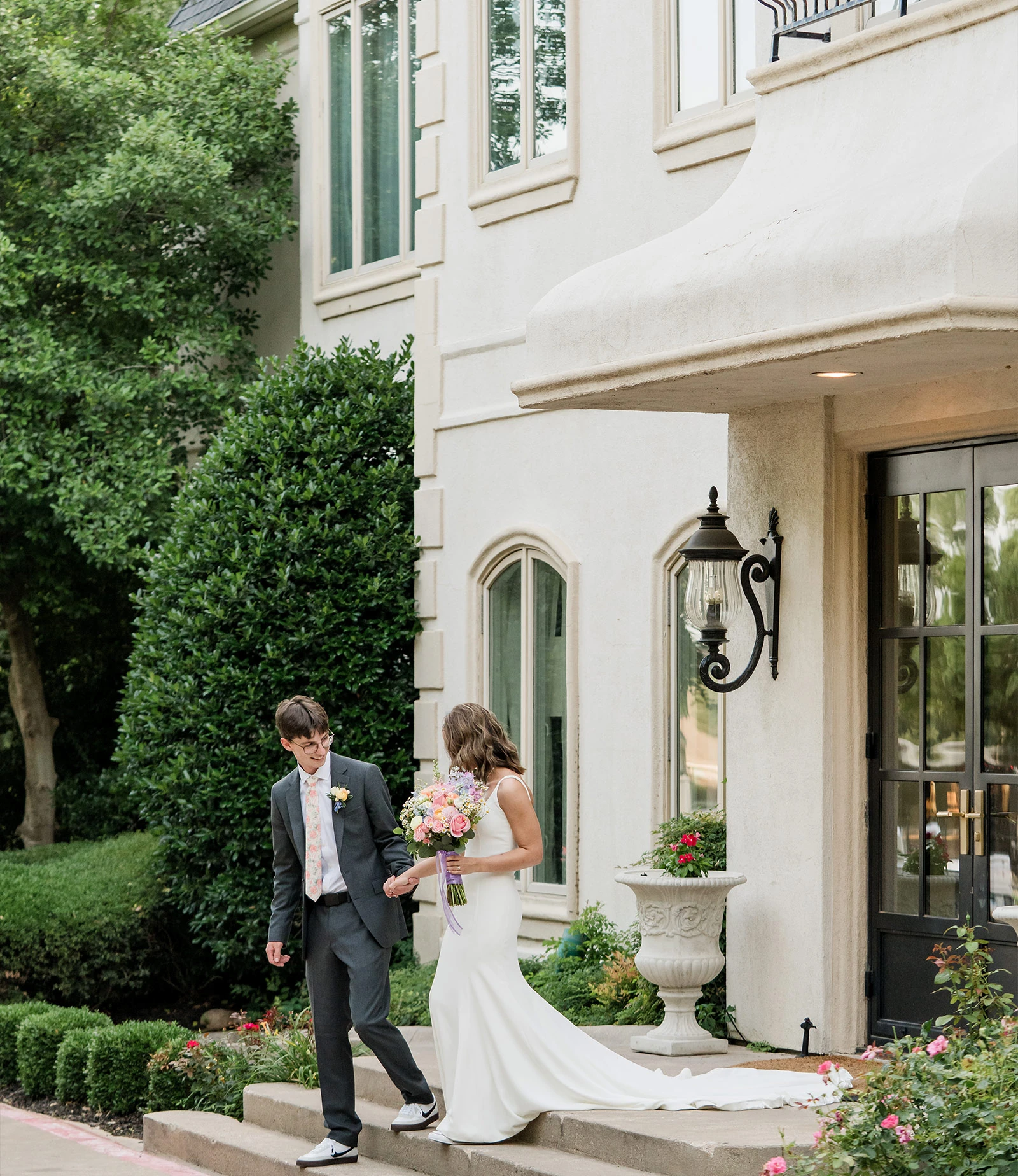 Outdoor wedding ceremony setting under a large, lush green tree with rows of white folding chairs arranged on a stone patio, and a white draped arbor with floral accents in the background