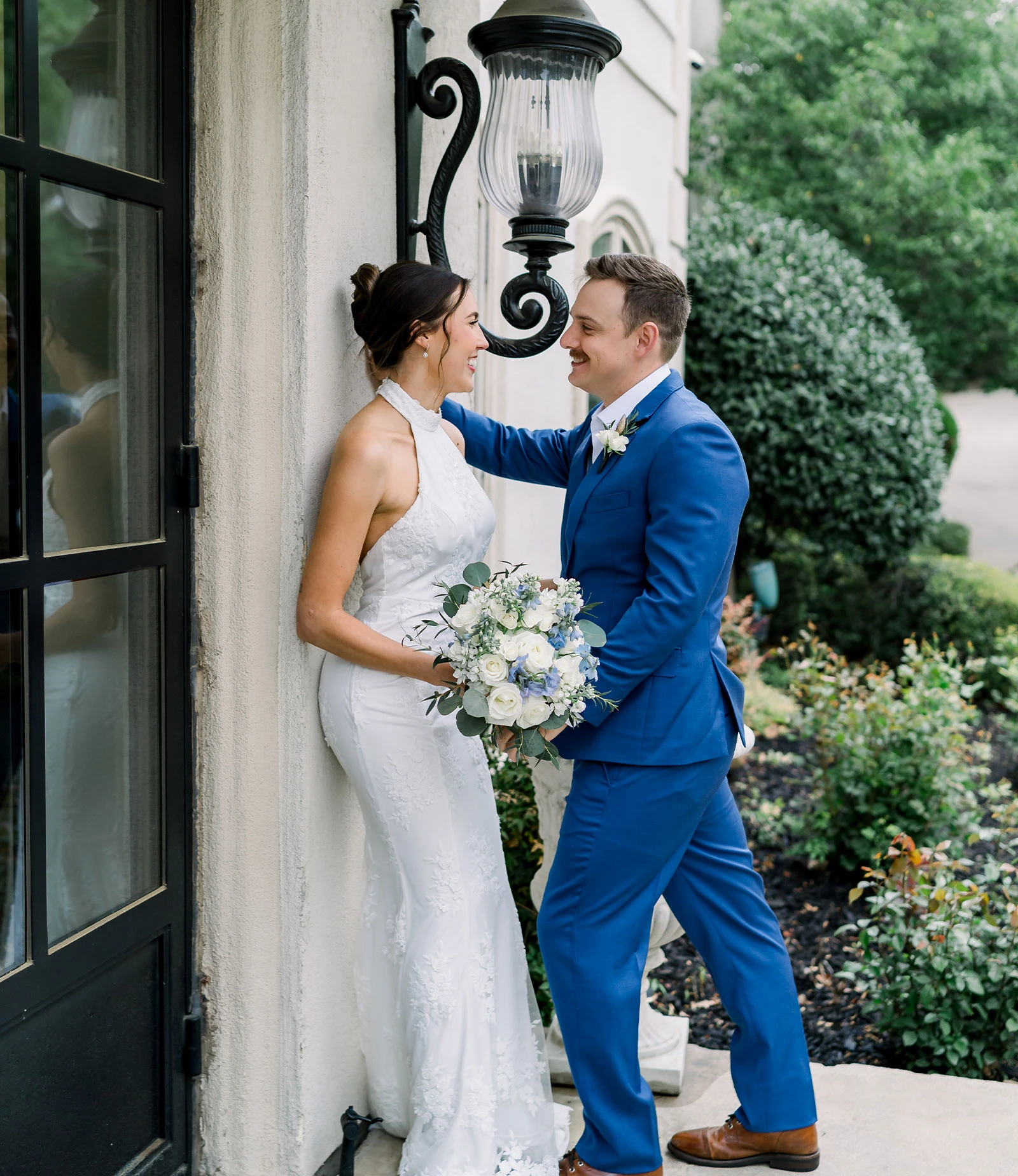 Two brides in white lace wedding gowns stand on the front steps of a white mansion, looking at each other, in front of a black double door entrance.