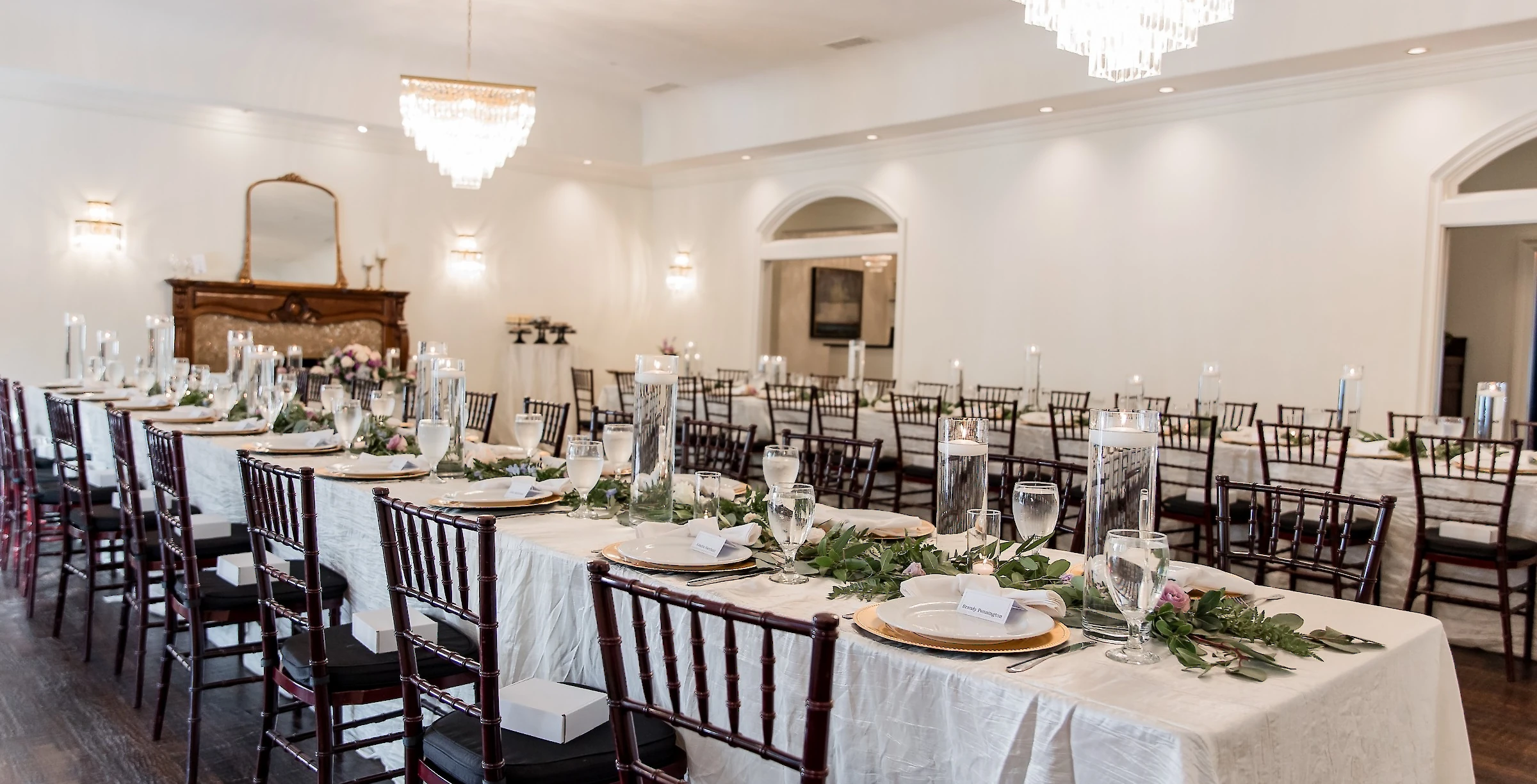 An elegant wedding reception hall featuring long tables draped in white linens, set with gold chargers, clear candles, and green garlands, under crystal chandeliers.