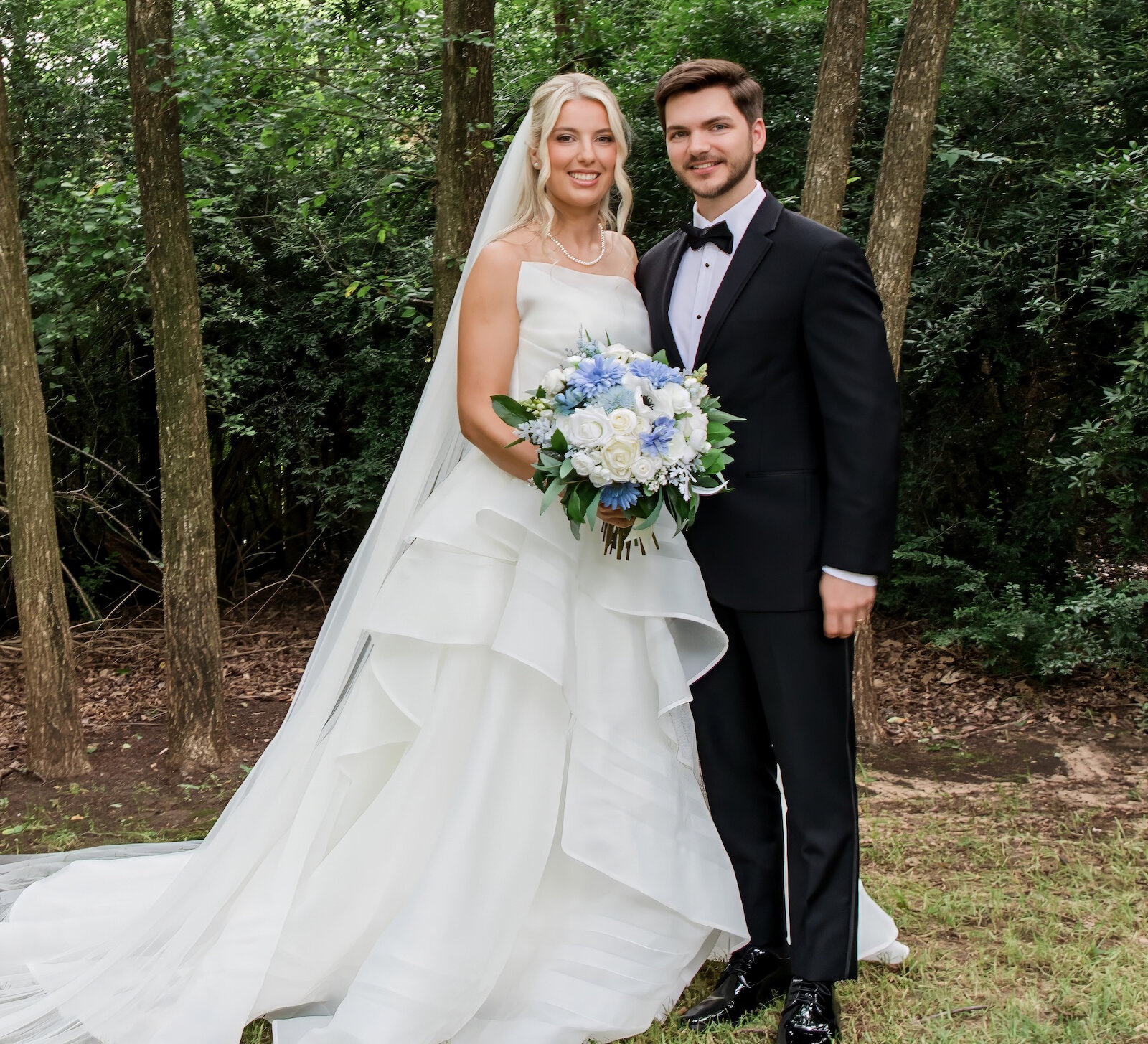 A beautiful full-length portrait of the bride and groom standing in a wooded area, with the bride holding a white and blue ruffled bouquet and wearing a layered gown.