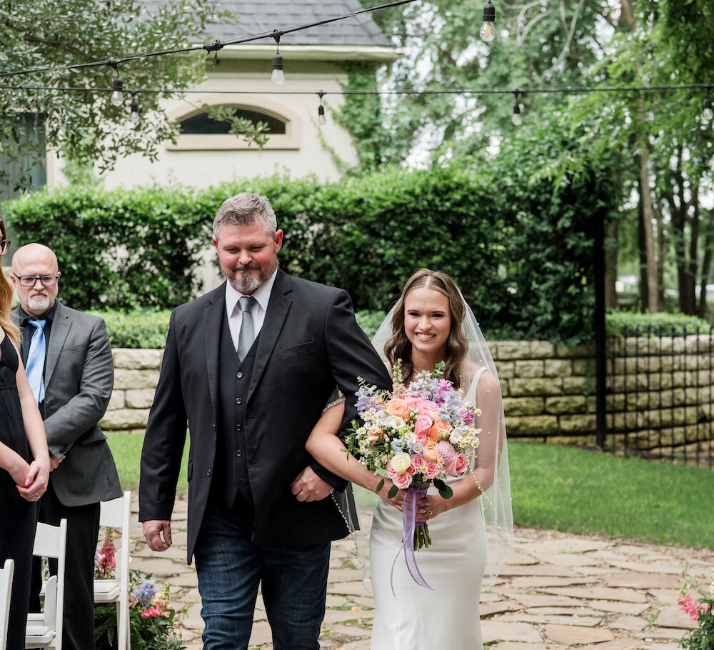 The smiling bride walks down the stone aisle arm-in-arm with her father, holding a large pastel and purple bouquet, during the outdoor ceremony procession.