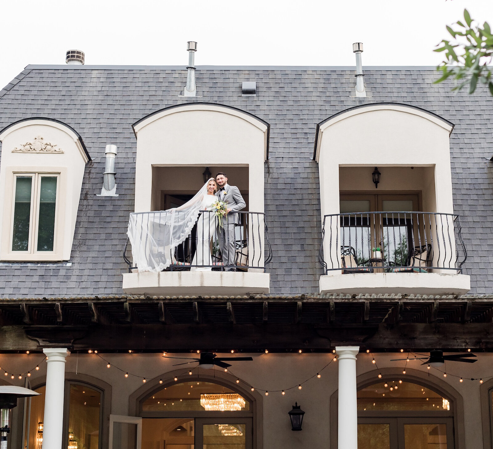 The bride and groom pose on an upper-level balcony of a mansion venue with a dark roof and dormer windows, with the bride's veil trailing in the breeze.