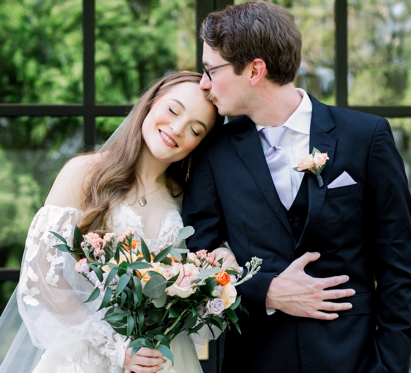 The groom in a black suit kisses the bride on the forehead as she holds a colorful bouquet, standing in front of black French doors with glass panes.