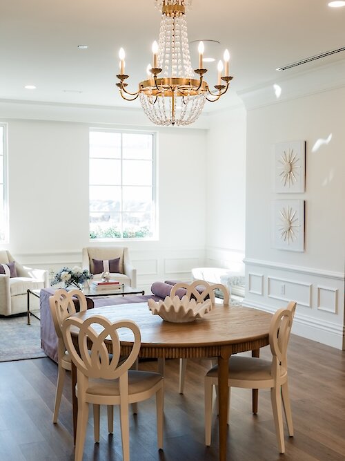A bright, modern interior room featuring a wooden circular table with unique chairs and a crystal gold chandelier hanging overhead.