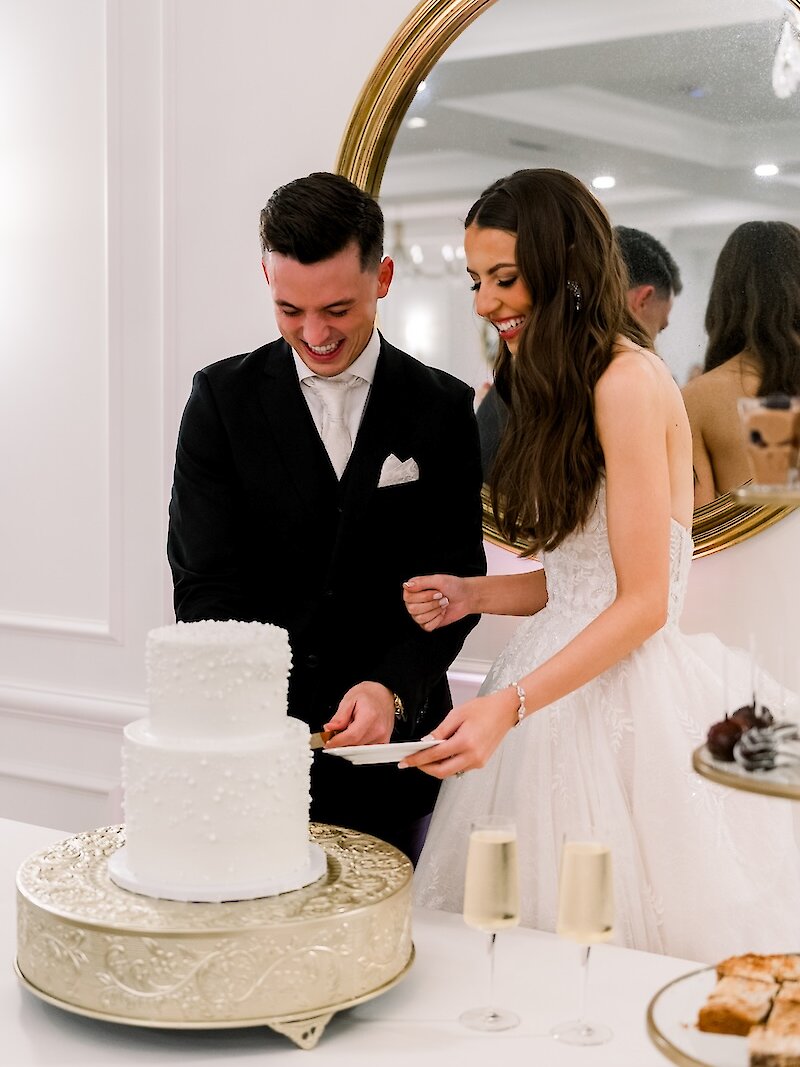 A bride and groom laugh together as they cut their white wedding cake during their reception.