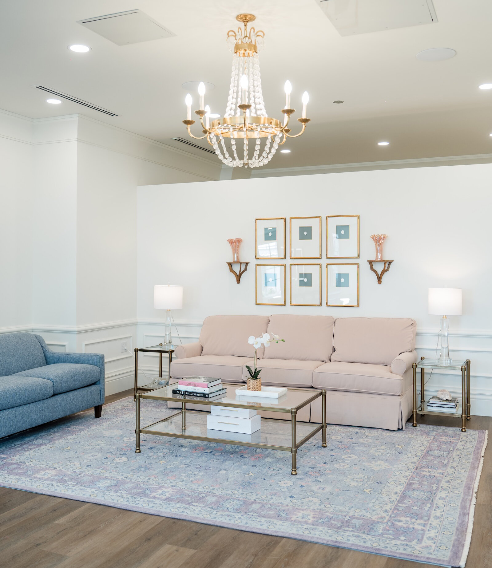 Elegant lounge room with a blush sofa, blue armchair, glass coffee table, pastel area rug, and gold chandelier in a bright, stylish interior.