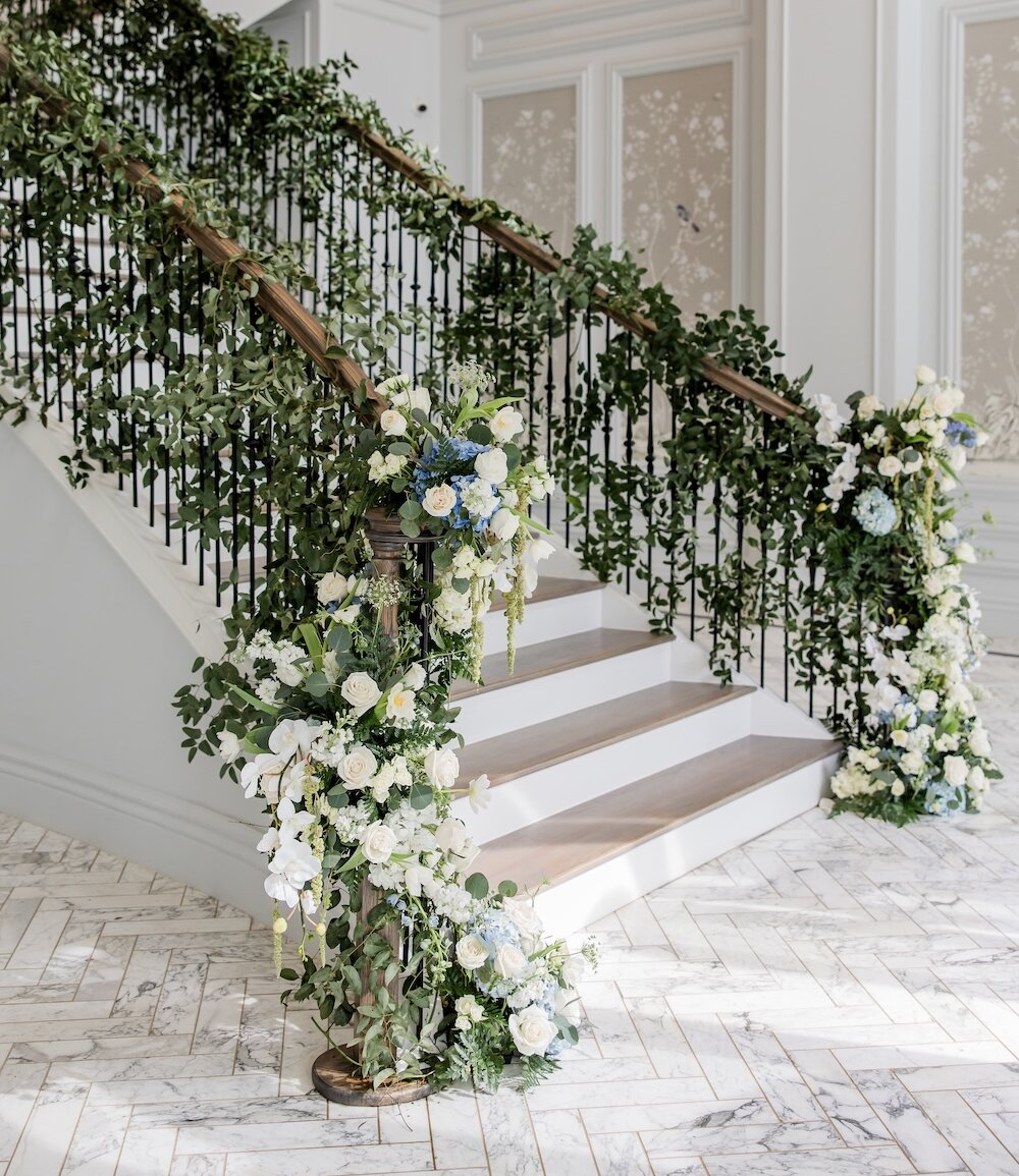 A grand white staircase with dark railings is heavily decorated with lush greenery garlands and clusters of white and blue flowers for an event.