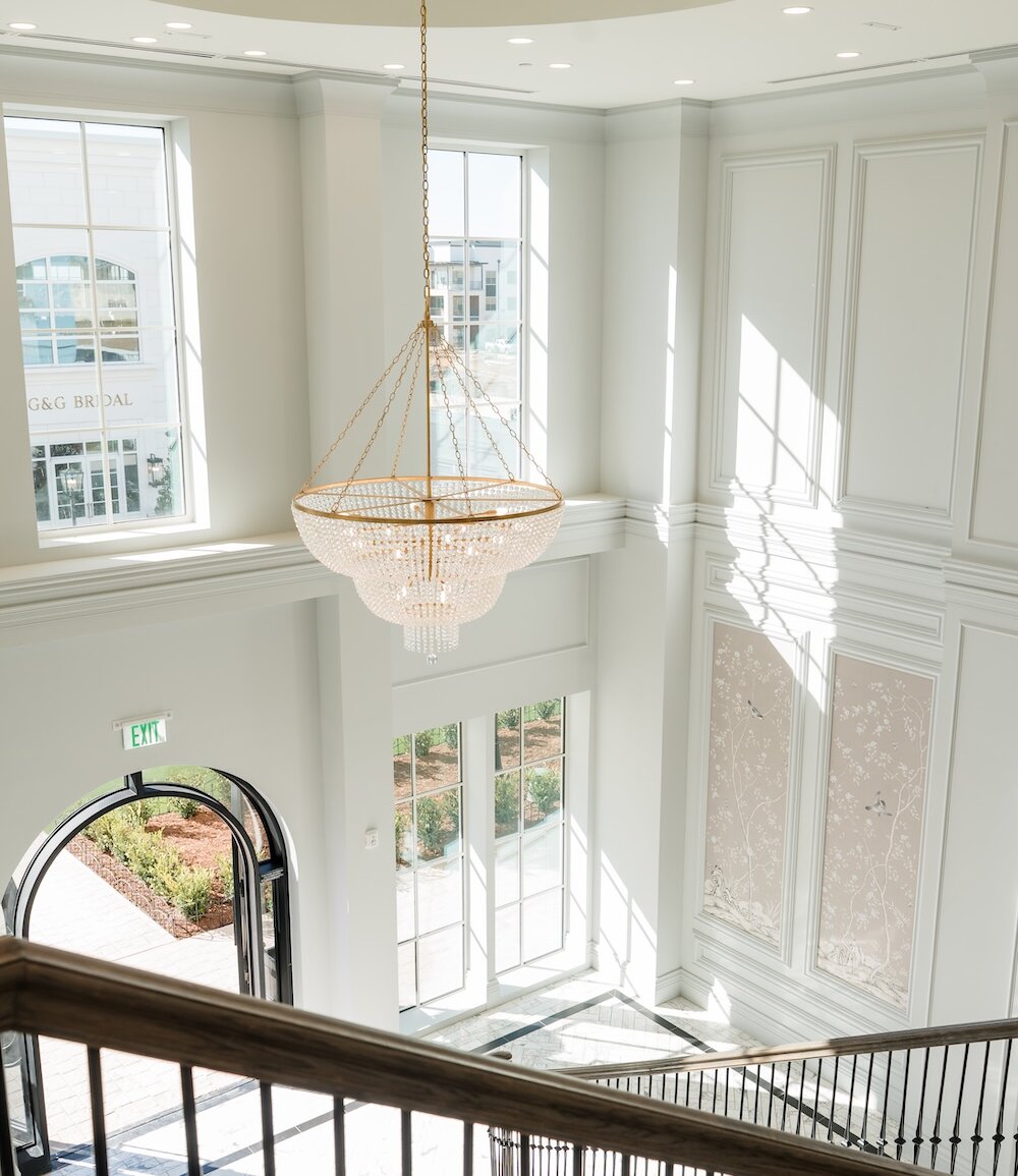 Grand staircase foyer filled with natural light, featuring tall windows, elegant wall paneling, and a large gold crystal chandelier.