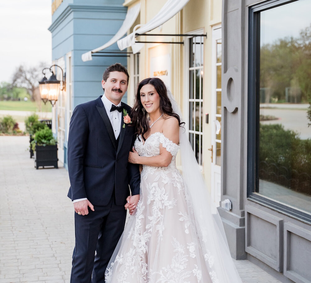 The bride in an off-the-shoulder lace gown holds hands with the groom in a navy tuxedo while posing outside a colorful building facade.