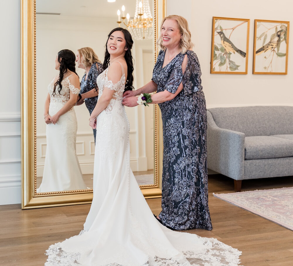 Mother helps the bride button her lace wedding gown as they smile in front of an ornate gold mirror.