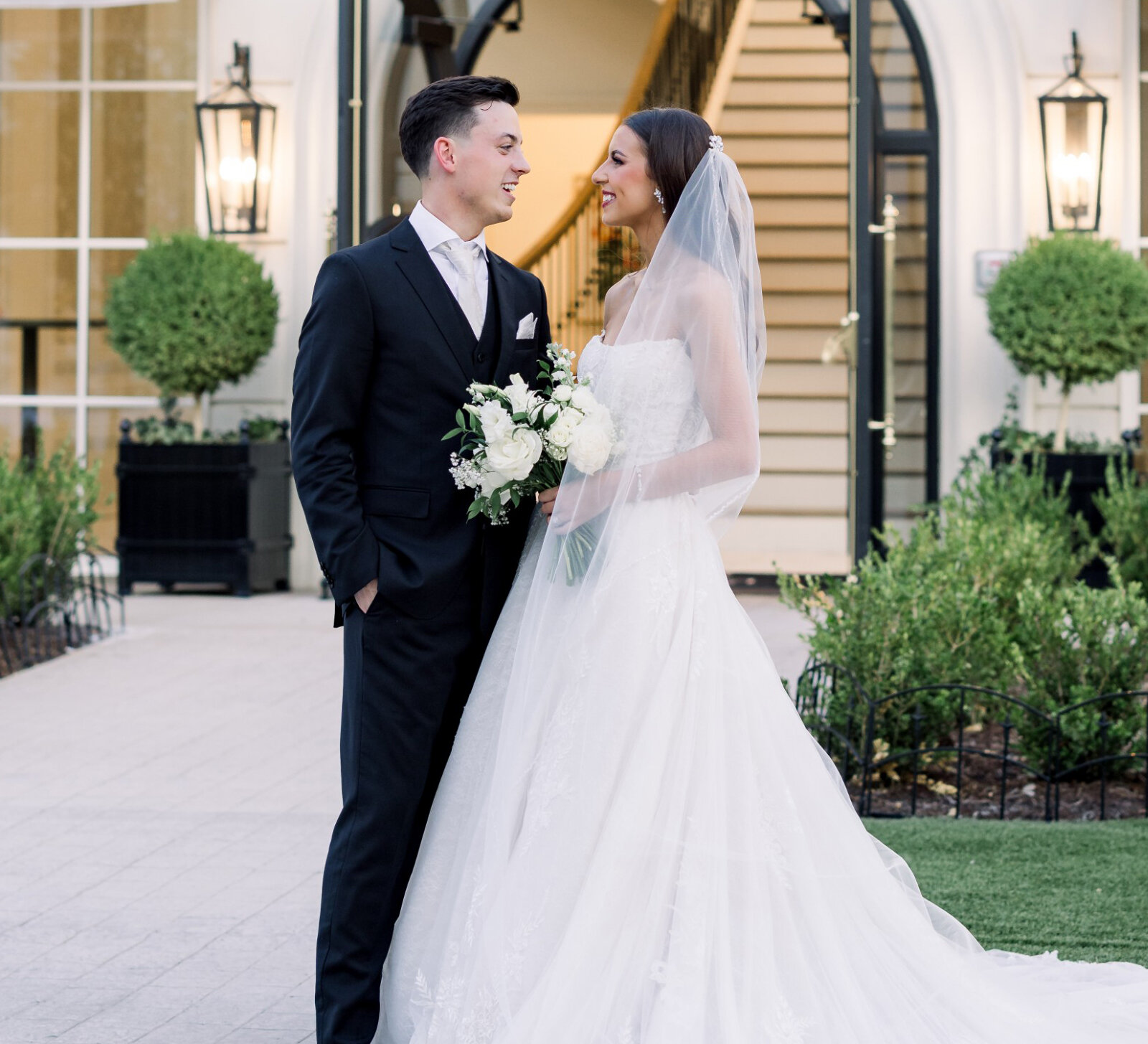 Bride and groom smiling at each other while standing outside a modern white venue, with the bride holding a bouquet and her long lace train spread across the grass.