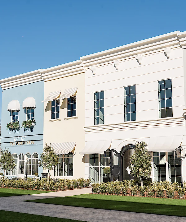 Colorful row of boutique-style buildings with blue, cream, and white facades, striped awnings, tall windows, and neatly landscaped greenery under a bright blue sky.