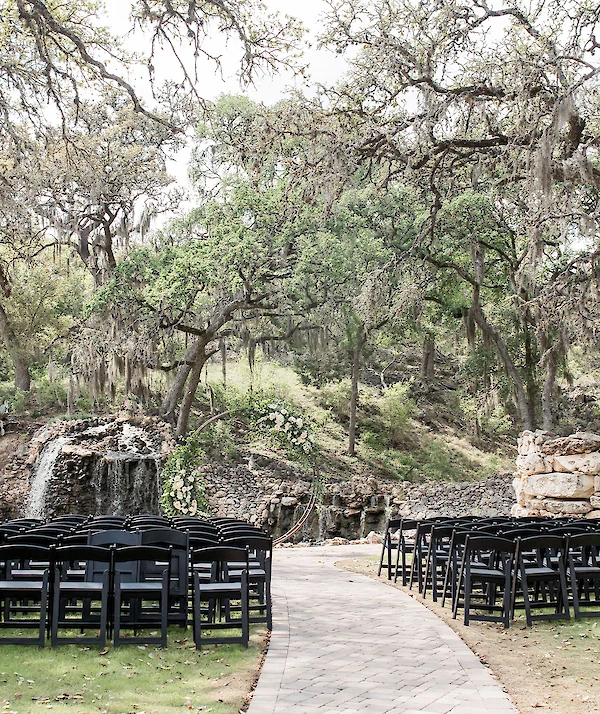 Modern white venue with gabled roofs, wood accents, large windows, and a covered entrance, surrounded by mature trees, stone landscaping, and a small footbridge.