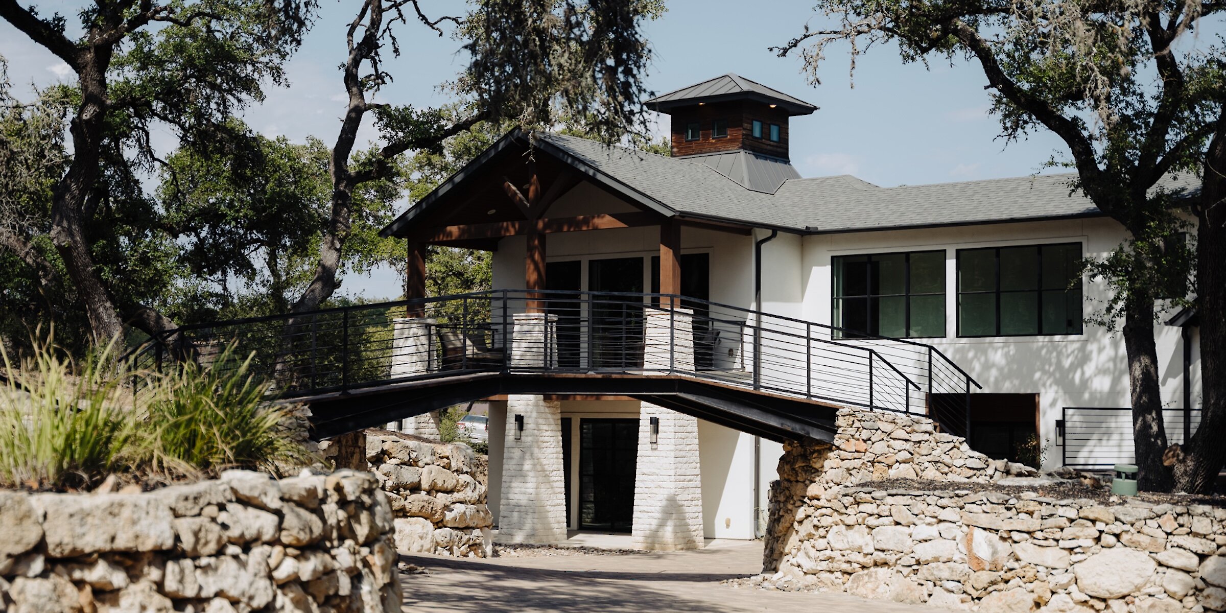 Modern wedding venue exterior featuring a white building with large windows, a wooden porch, and a raised black metal bridge supported by stone walls, surrounded by tall oak trees and natural landscaping.