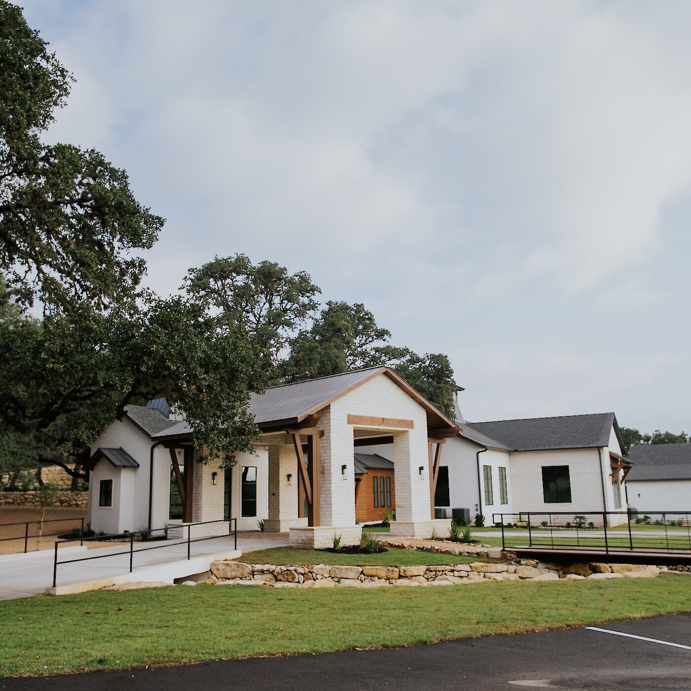 Modern white wedding venue exterior with stone accents, oak trees, and a small bridge over landscaped grounds