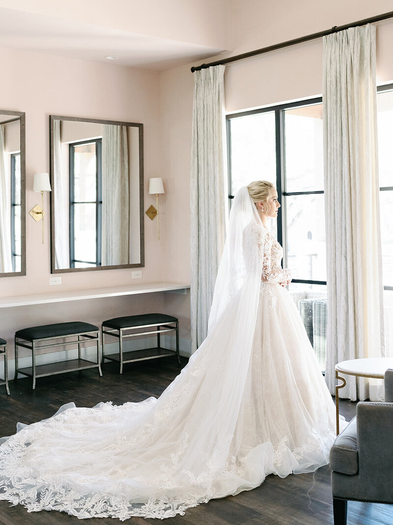 Bride with long, cascading braids sits gracefully by a large window, holding a romantic bouquet of pink and white flowers while wearing a flowing wedding gown with a long train.