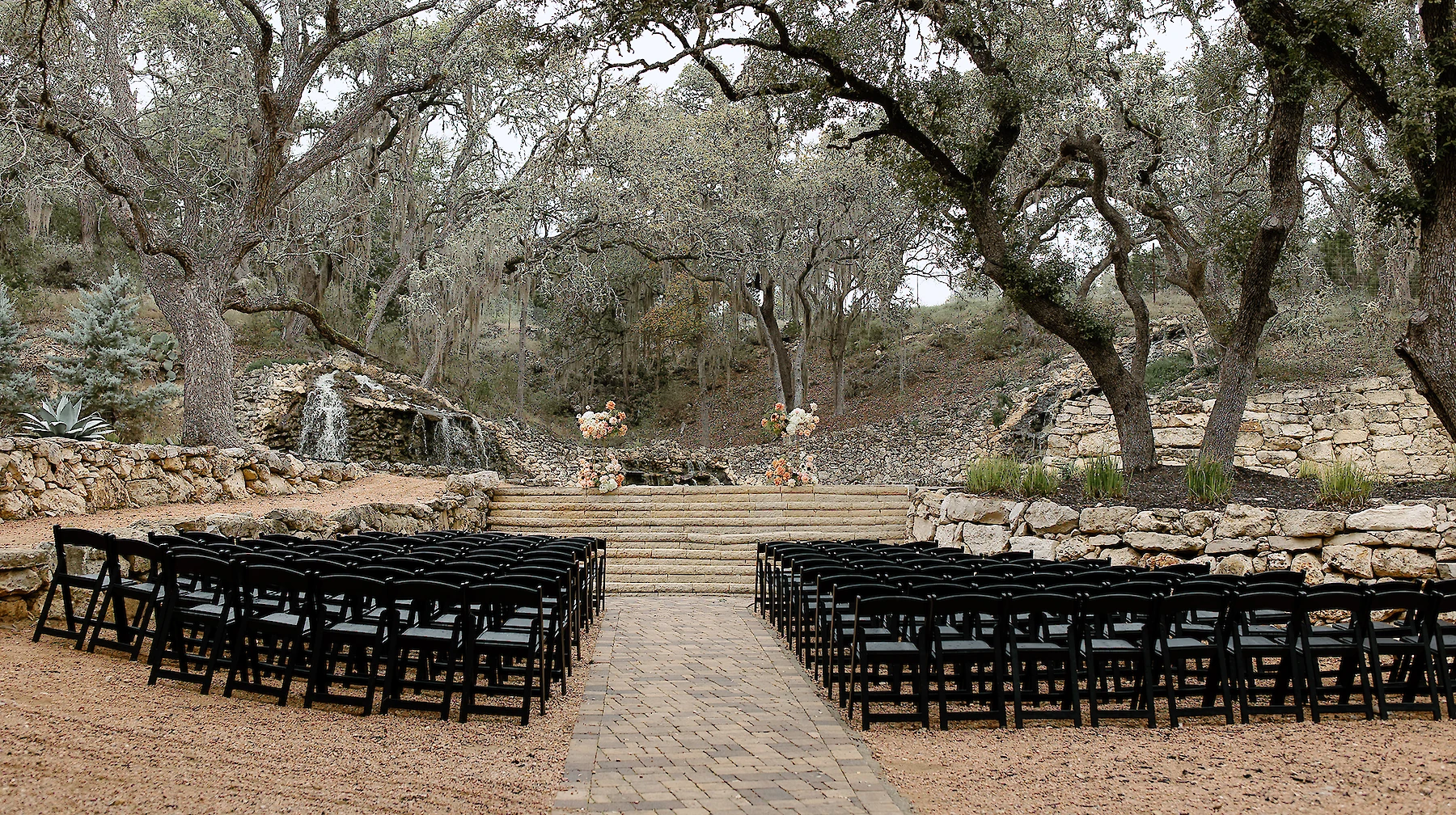 Outdoor wedding ceremony setup featuring rows of black chairs facing stone steps and a cascading waterfall backdrop, surrounded by large oak trees and natural stone landscaping.