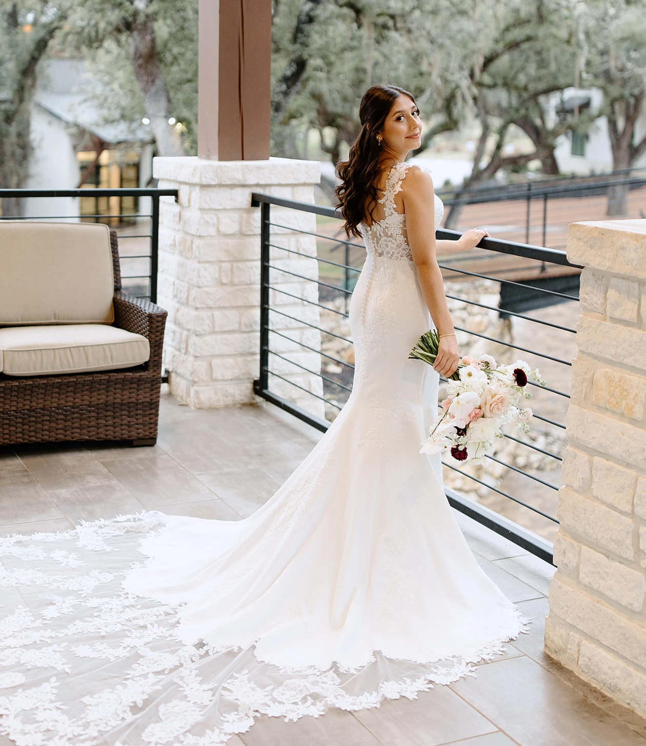 Bride in a fitted lace-back wedding gown with a long lace train, standing on an outdoor terrace and holding a bouquet while looking over her shoulder