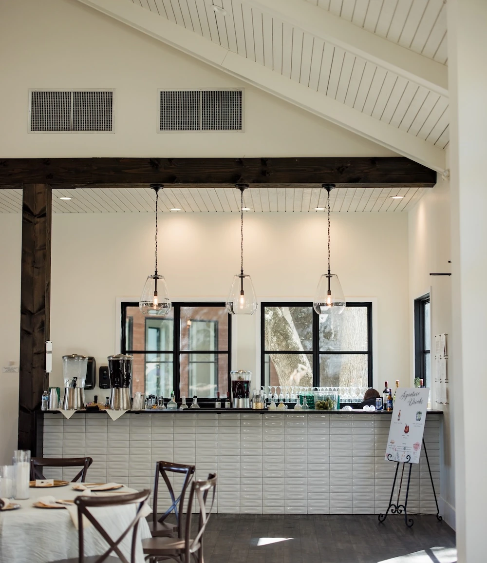 A modern farmhouse wedding venue bar area featuring white tiled walls, pendant lights, dark wood beams, and a signature cocktail sign.