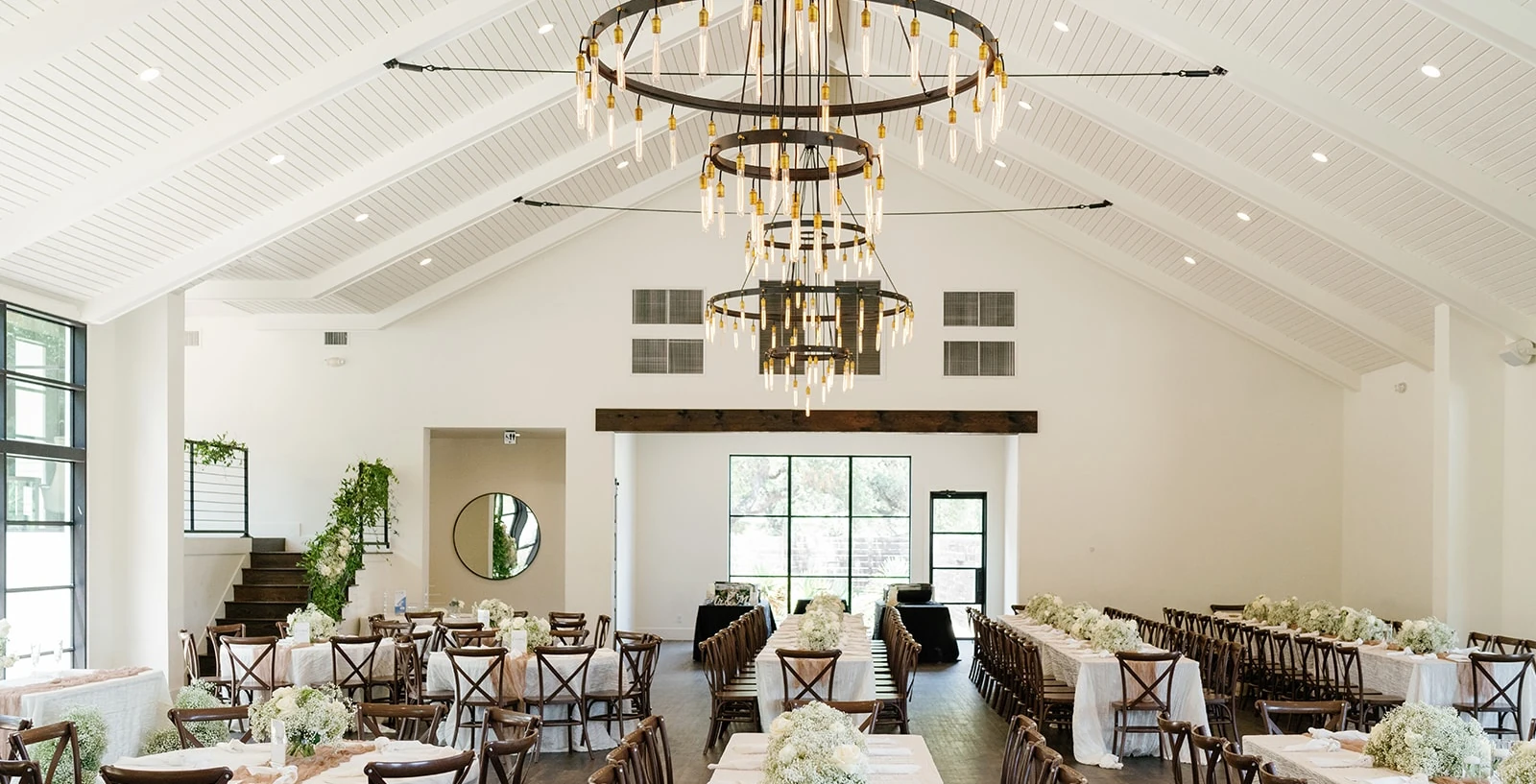 A bright, modern reception hall featuring a vaulted white ceiling, long dining tables, cross-back chairs, and large tiered ring chandeliers.