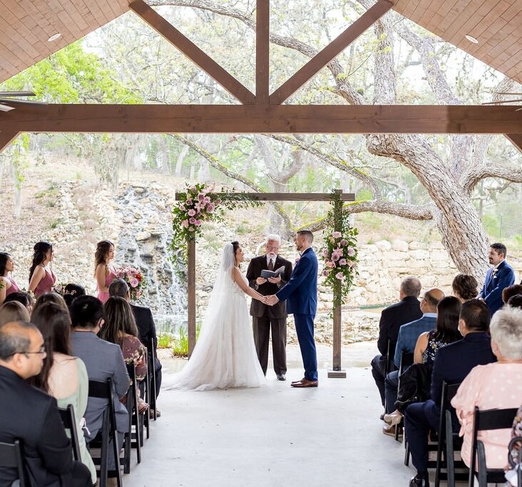 Outdoor pavilion wedding ceremony with bride and groom holding hands under pink floral arch, guests seated, stone waterfall backdrop, hill country venue.