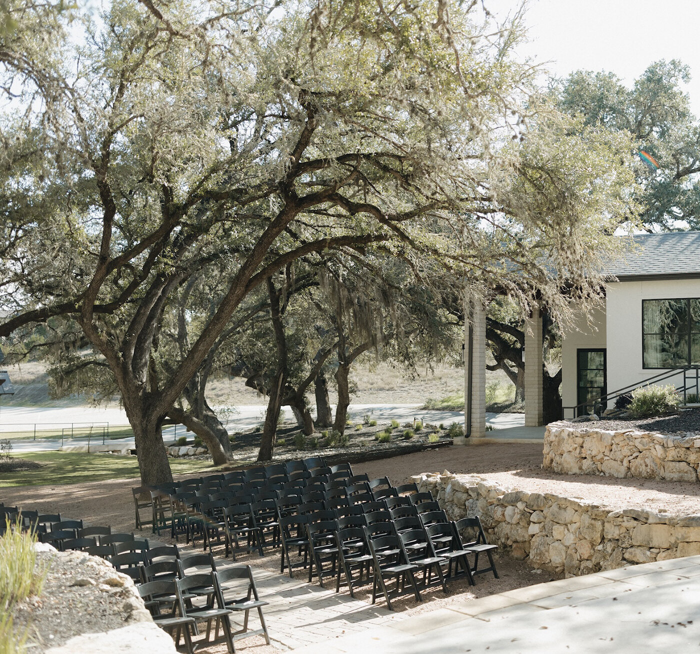 Outdoor wedding ceremony setup with rows of black chairs arranged beneath large oak trees beside a modern venue building