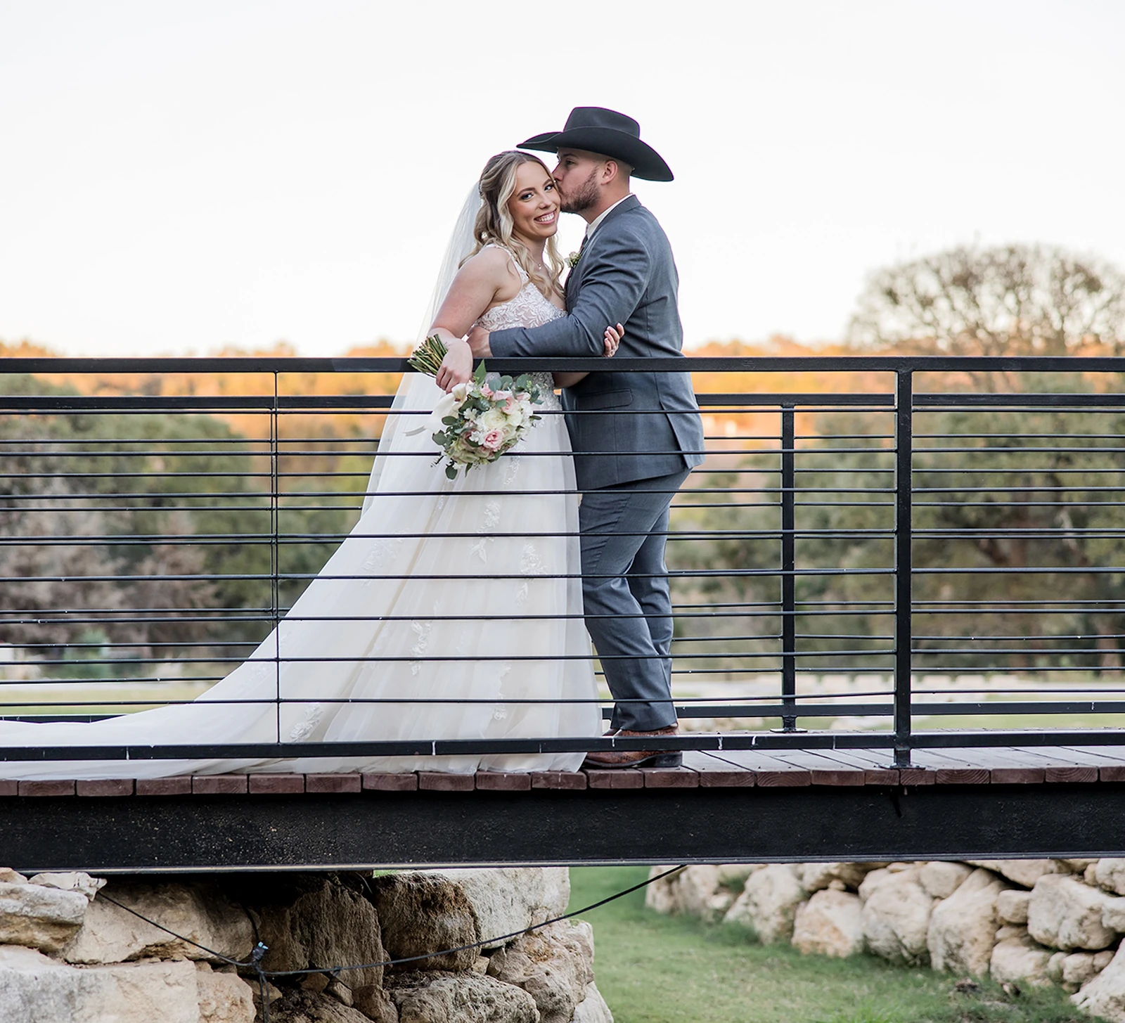 Bride and groom embrace on a rustic stone bridge, with the groom wearing a cowboy hat as they share a sweet moment at sunset in a scenic outdoor setting.