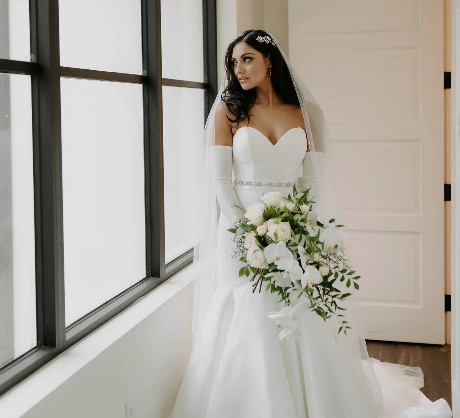 Elegant bride in a classic white wedding gown and veil standing by a window, holding a cascading bouquet of white roses and orchids