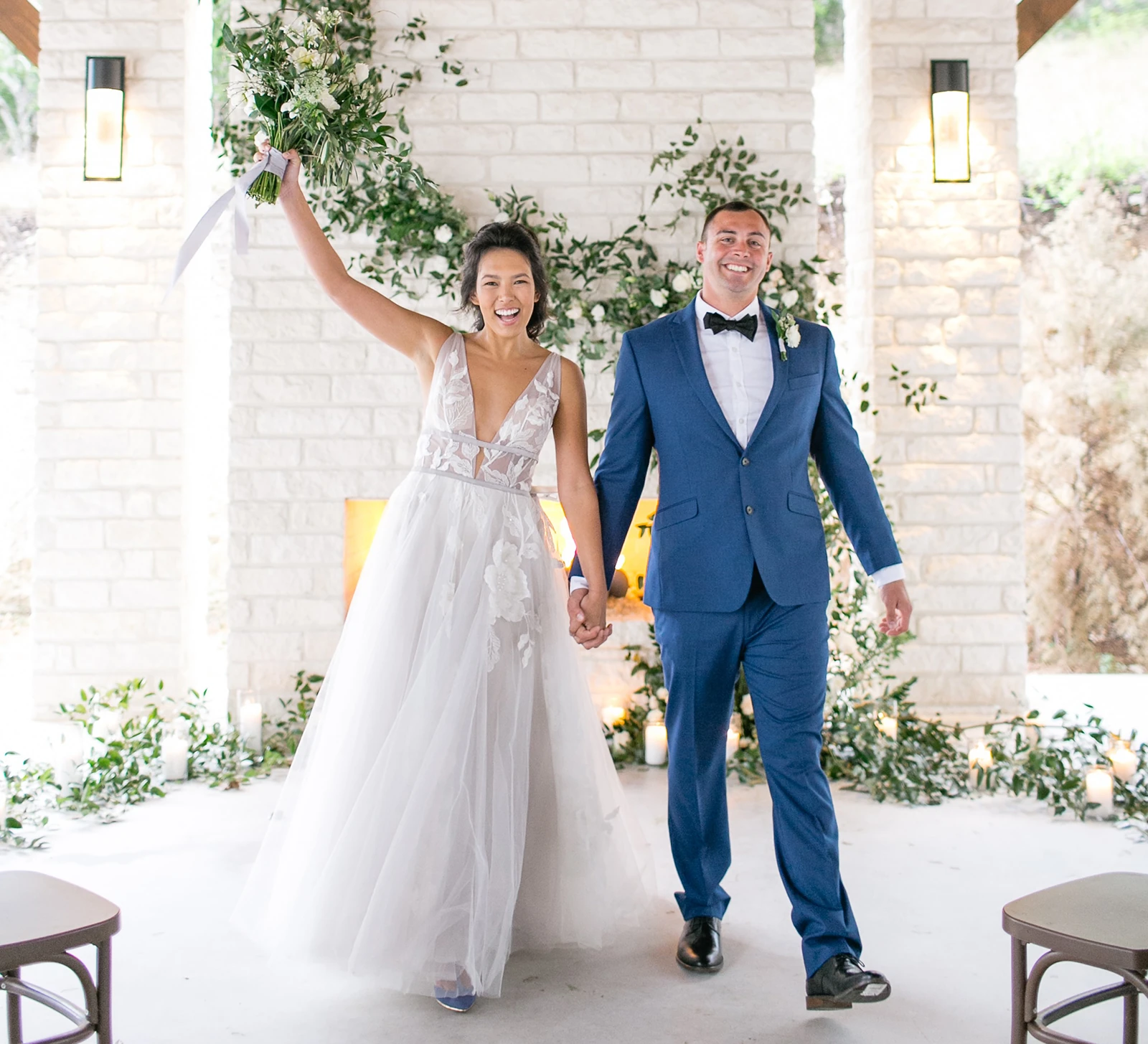 Bride and groom celebrating their wedding recessional under a covered outdoor pavilion with white stone fireplace and greenery décor.