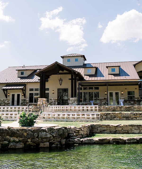 Large rustic-style venue with a metal roof, stone accents, covered patio areas, and tiered stone landscaping overlooking a pond under a bright sky.