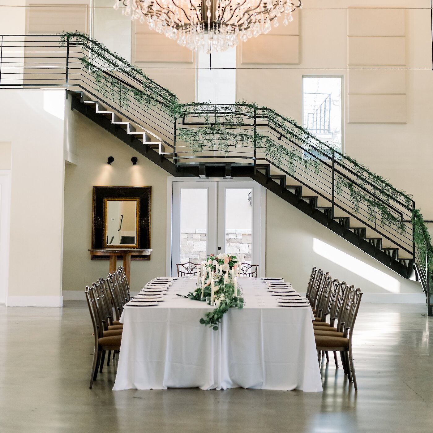 Elegant wedding reception hall with a long white-covered table, greenery centerpiece, tall candles, staircase draped in foliage, and a large crystal chandelier overhead.