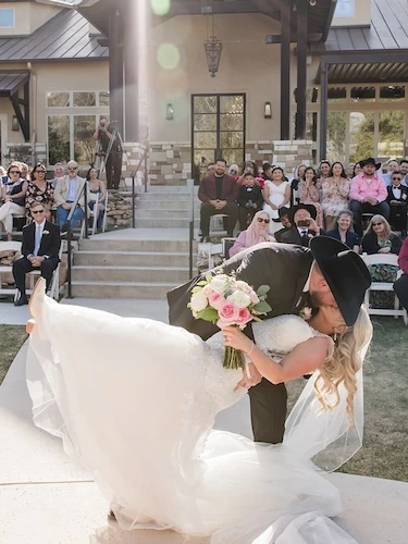 Bride and groom kissing in a dramatic dip during an outdoor wedding ceremony, surrounded by seated guests in front of a stone and metal-roofed venue.