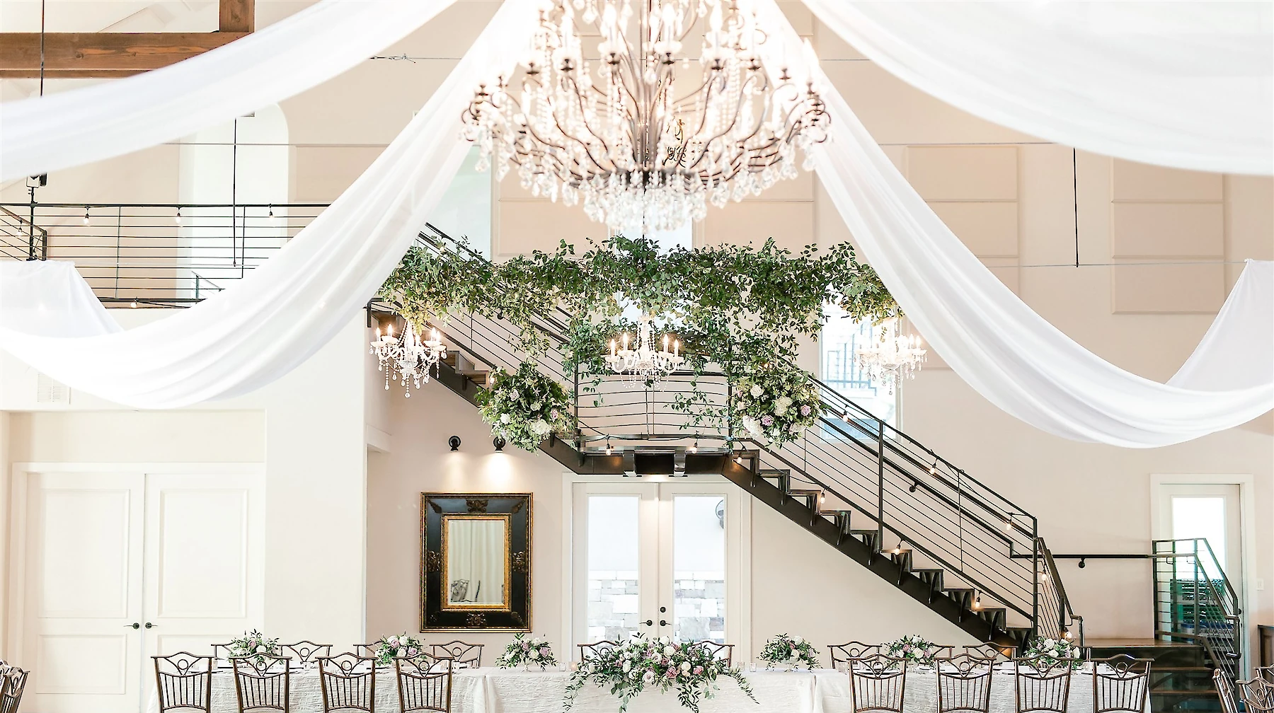 A luxurious wedding head table set against a staircase, featuring abundant white drapery, an overhead greenery installation, and a large crystal chandelier.
