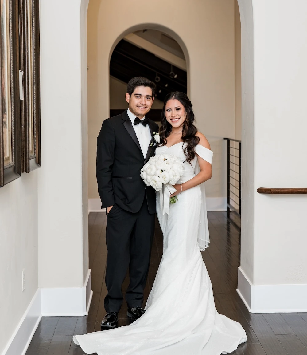 Bride and groom standing together under an arched hallway, smiling and holding a white floral bouquet.