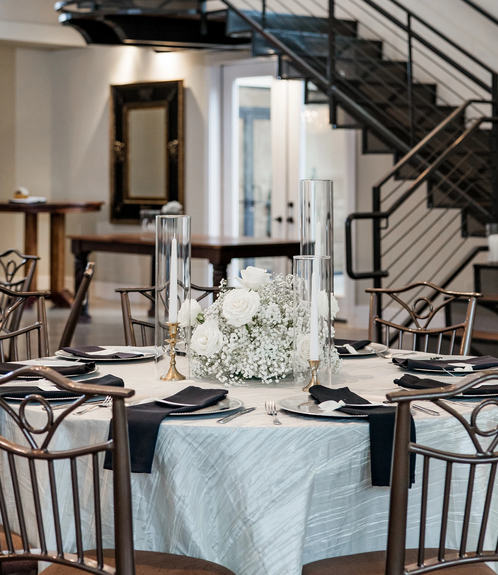Elegant wedding reception table decorated with white roses, baby’s breath centerpiece, black napkins, and tall glass candles in a modern venue with a staircase