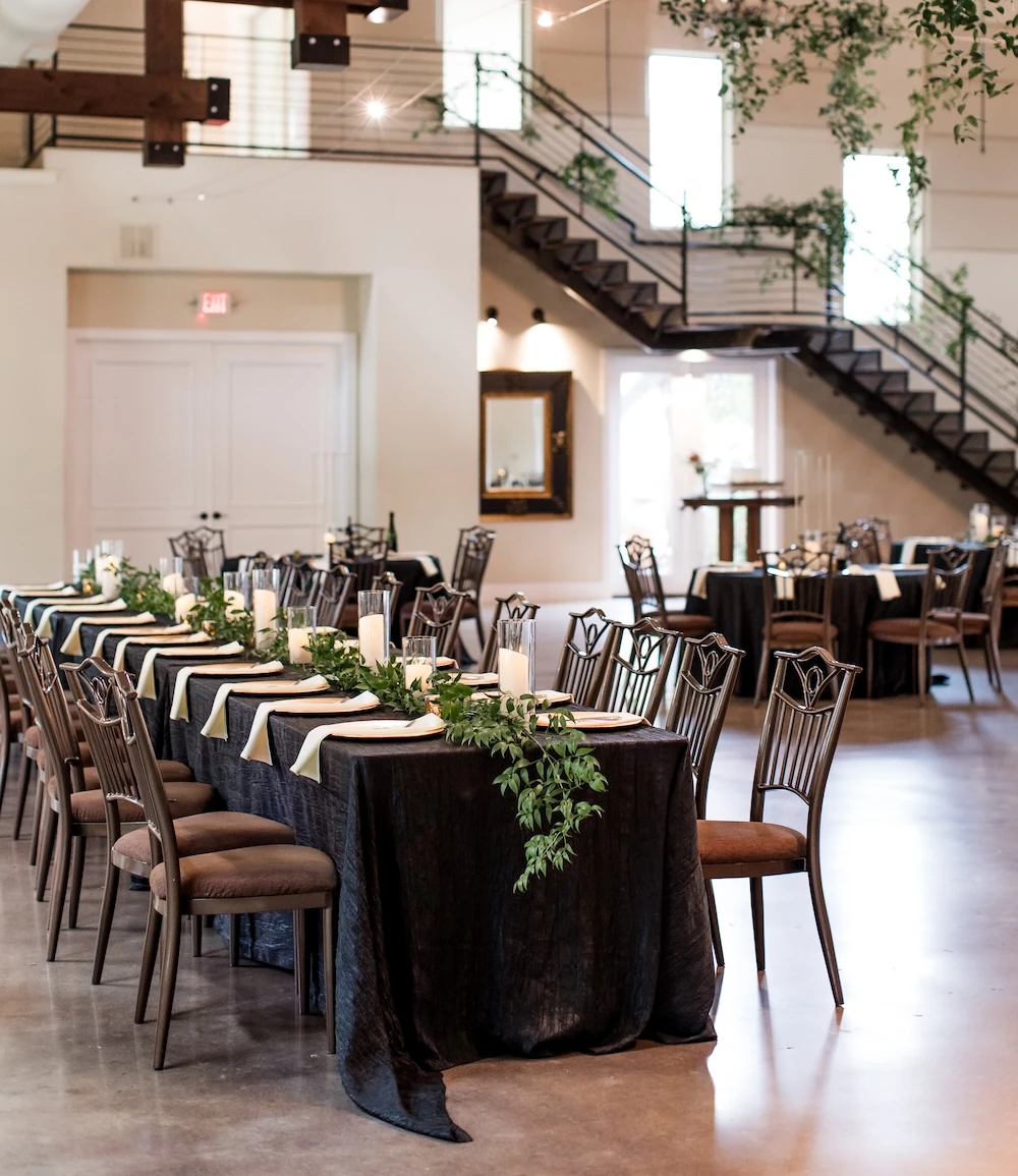 Elegant wedding reception setup inside modern venue with black linens, greenery centerpieces, and wooden beams overhead.
