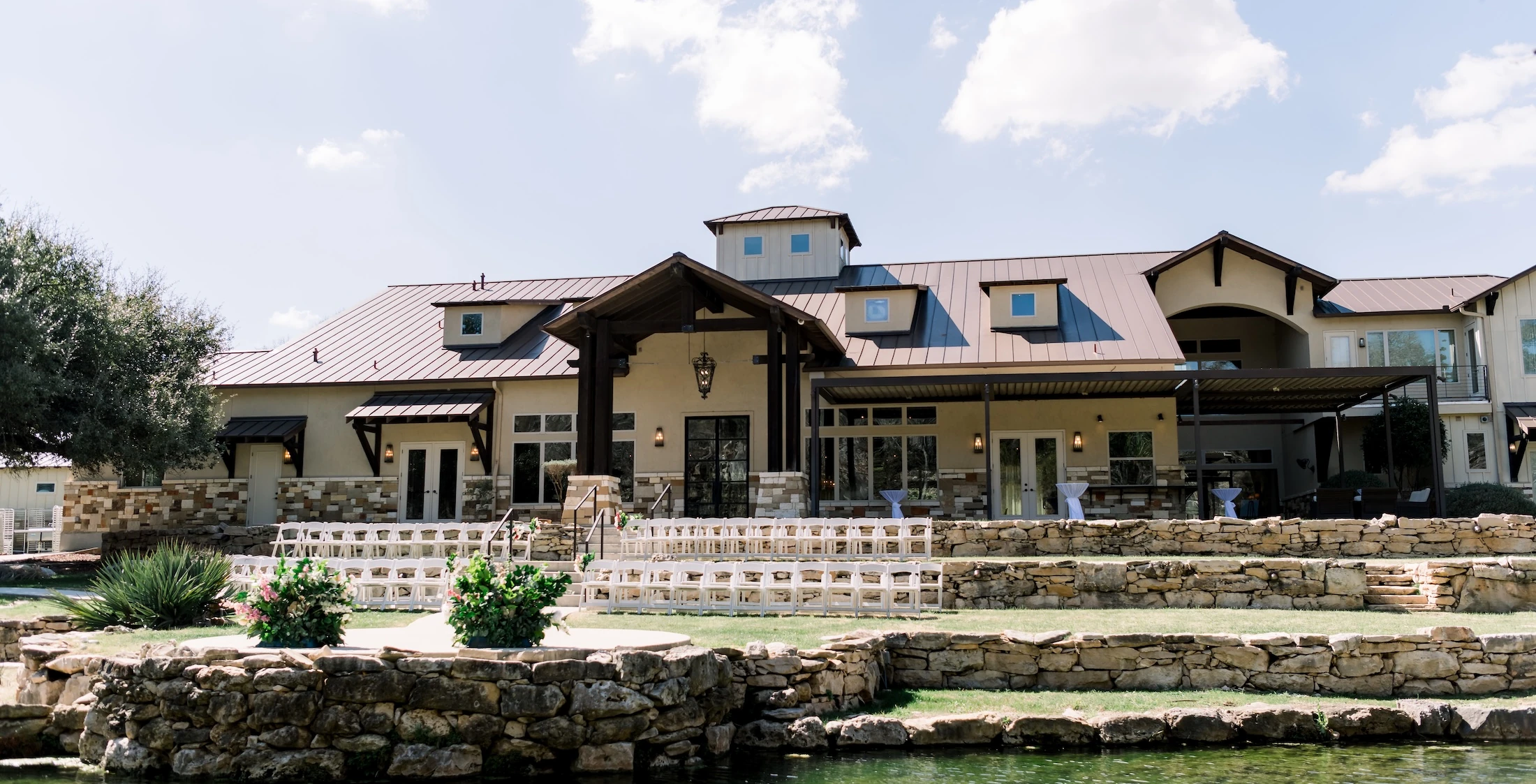 Outdoor wedding venue with tiered stone landscaping, clear pond water, and a large modern lodge featuring a metal roof and ceremony seating arranged on the lawn.