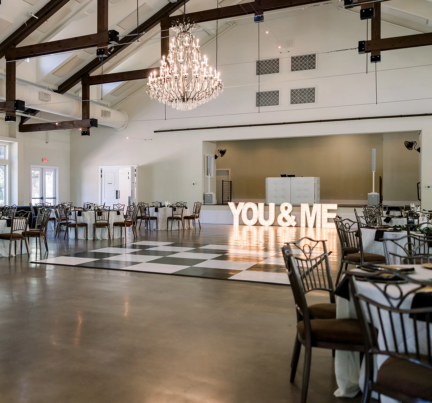 Spacious wedding reception hall featuring a black and white checkered dance floor, large ‘YOU & ME’ marquee letters, elegant chandeliers, and round guest tables