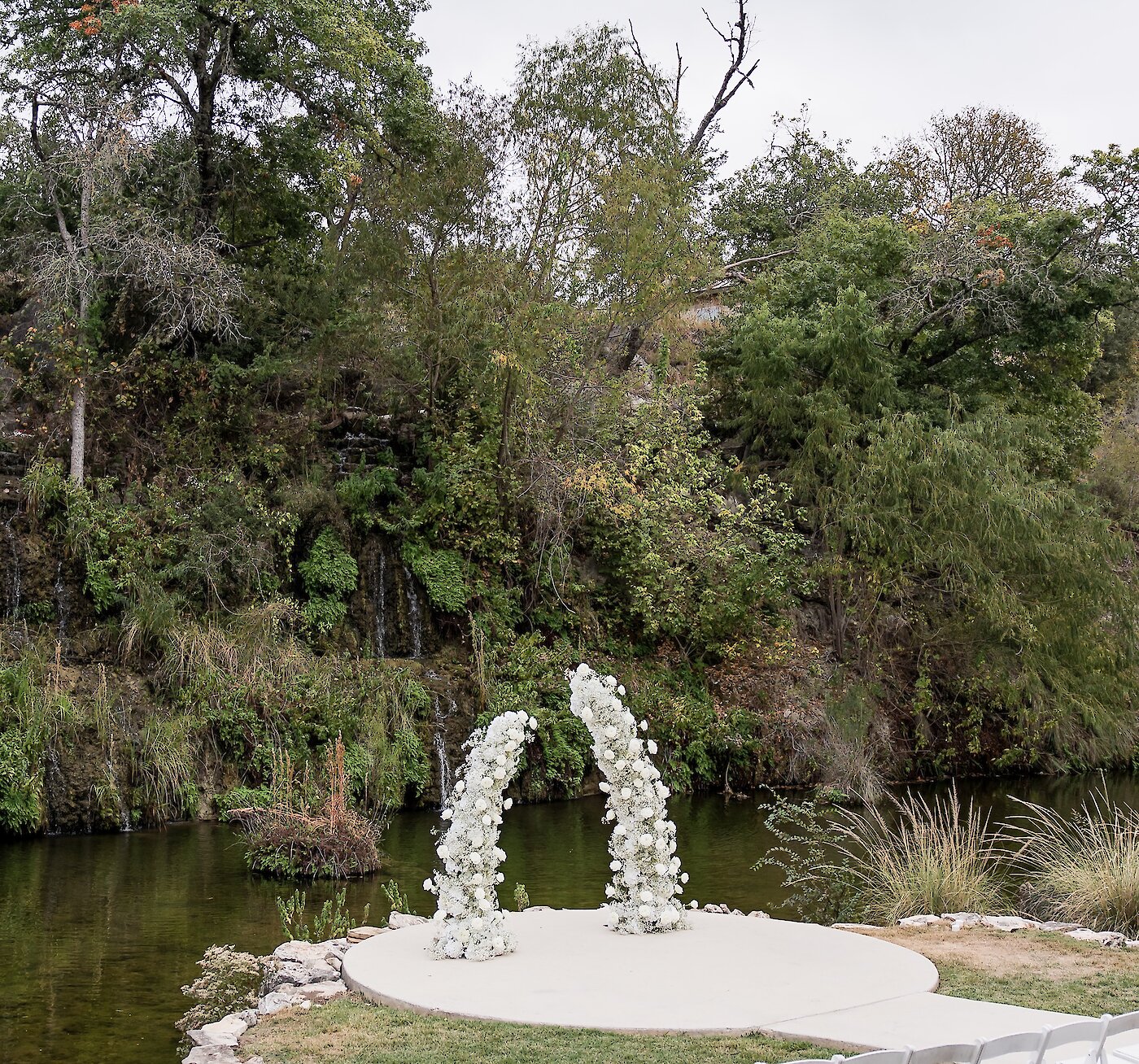 An outdoor wedding ceremony site with rows of white chairs facing a white floral arch on a round platform next to a pond and natural waterfall.