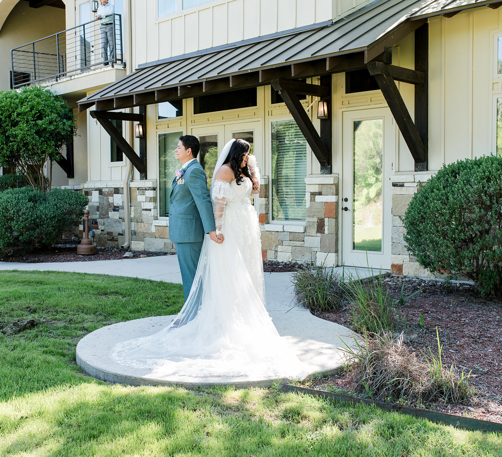 Bride and groom stand back-to-back holding hands outdoors before their first look at a wedding venue.
