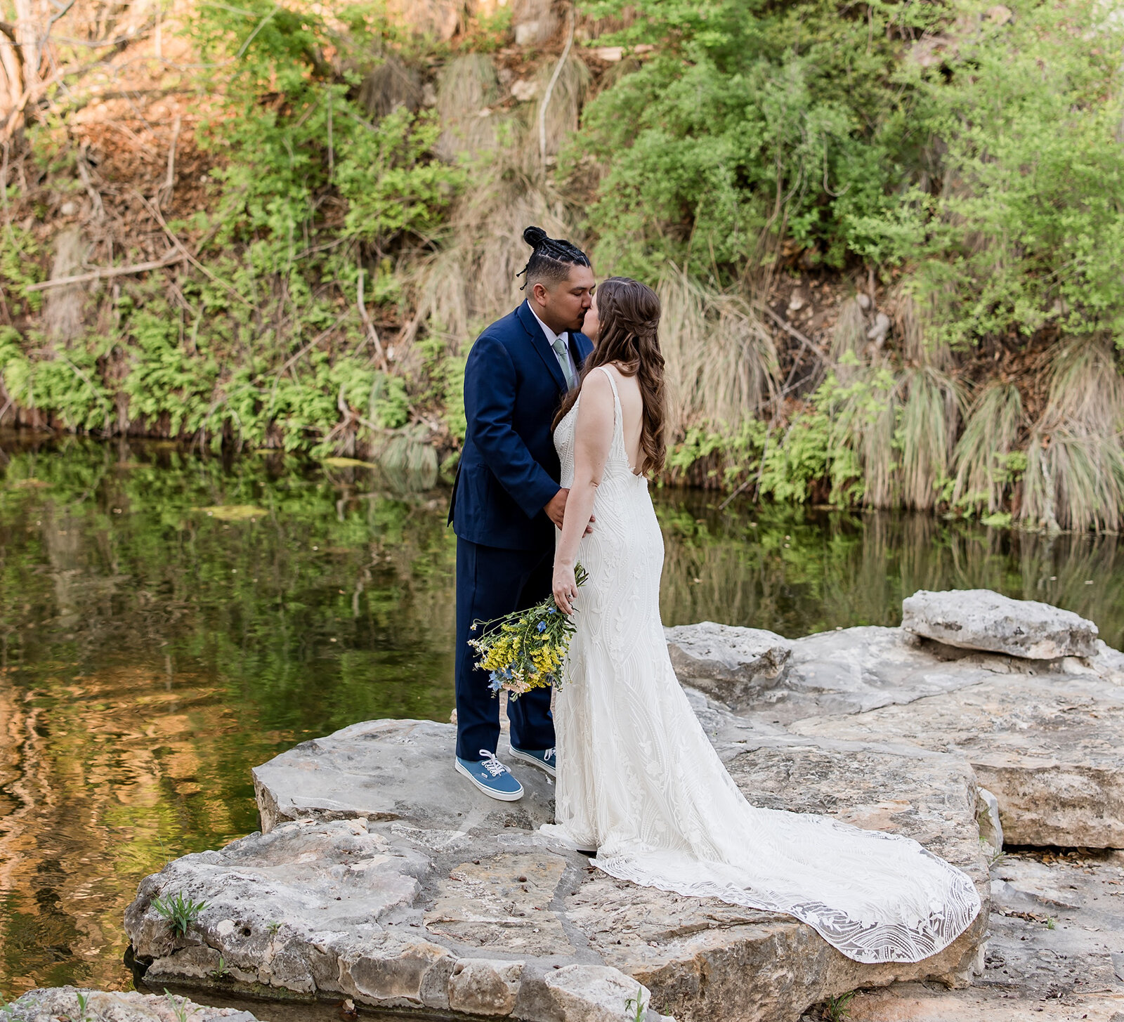 Bride and groom kissing on rocky ledge beside a peaceful creek surrounded by lush greenery at an outdoor wedding.