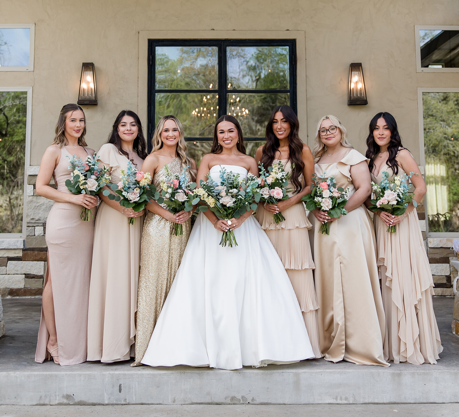 Bride standing with six bridesmaids in neutral and gold dresses, all holding floral bouquets during an outdoor wedding portrait